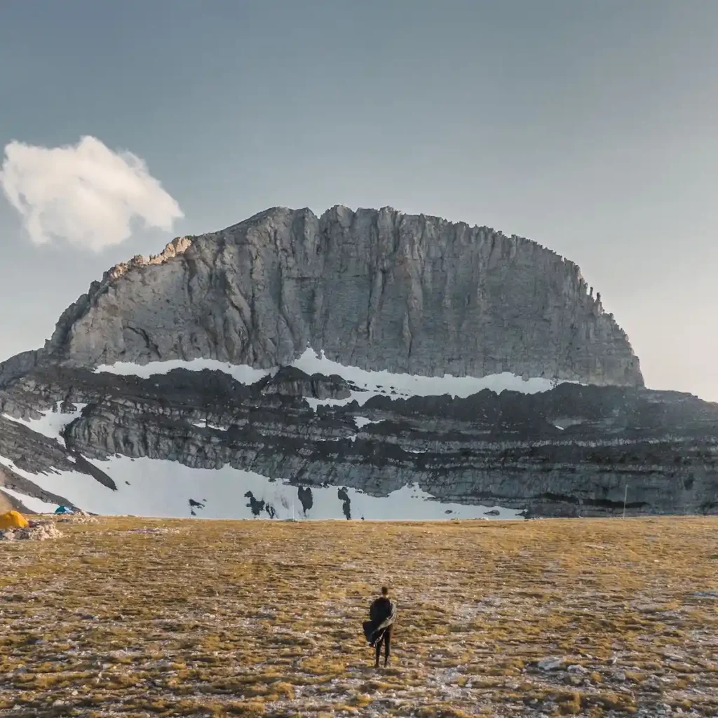 A lone hiker standing on a rocky mountain trail looking toward the soaring crags and peaks of Mount Olympus under a clear sky.