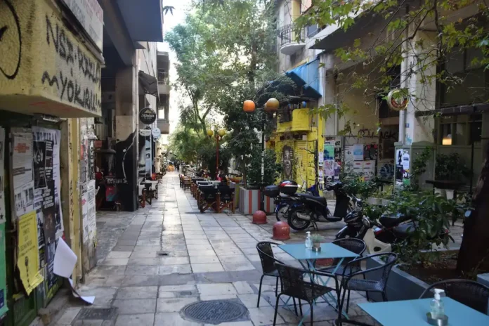 A pedestrian street in Exarcheia with café tables set outside, motorbikes parked along the curb, and walls covered in murals, tags, and posters.