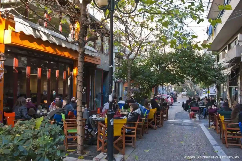 A pedestrian street in Exarcheia lined with cafés, where groups of people sit outdoors talking and drinking coffee.