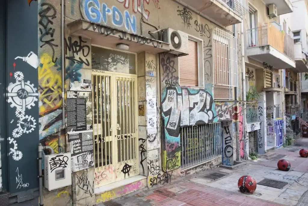 Street-level view in Exarcheia showing a condominium entrance door covered in graffiti tags, walls layered with political posters, and markings extending onto the sidewalk itself.