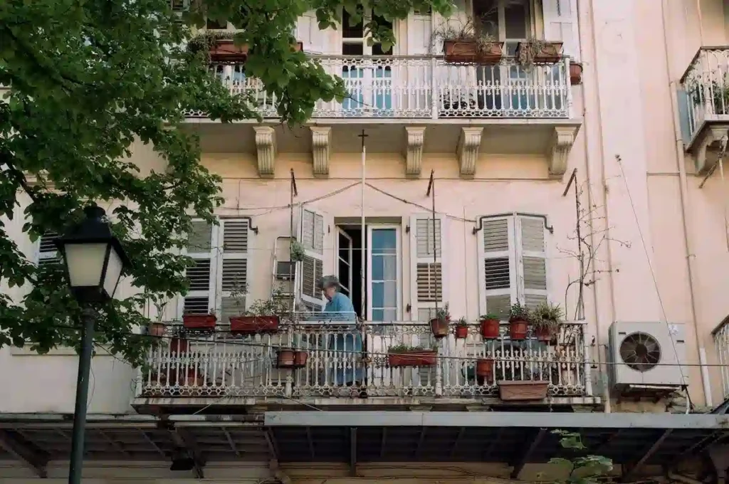 An elderly woman standing on a traditional balcony overlooking a historic street in Corfu Town