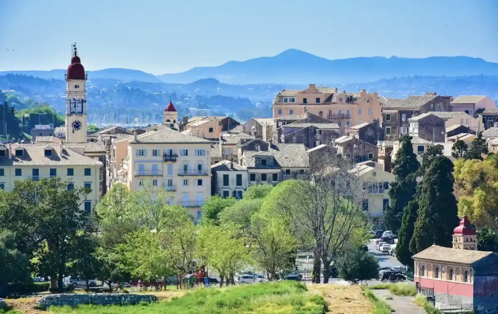 Green park in the foreground with a clock tower church, terracotta roofs, and big mountains in the background