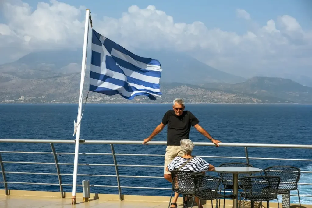 A man standing and a woman sitting on the open deck of a Greek ferry boat at sea.