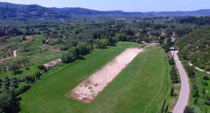 Aerial view of the ancient grassy stadium in Olympia, framed by olive trees and historic archaeological ruins.