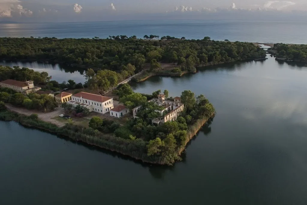 Aerial view of the Kaiafas thermal spring facilities nestled on an island in the lake, surrounded by pine forests and wetlands.