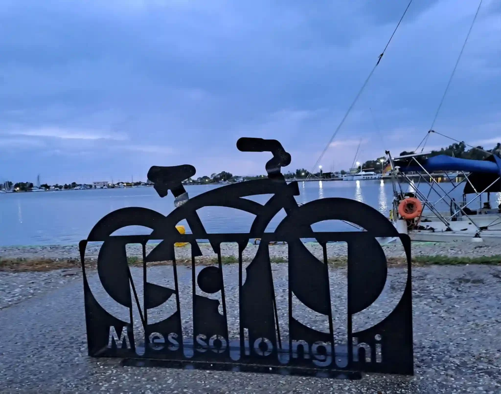 A sculptural bicycle stand shaped like a bike, with a Messolonghi sign and the town's lagoon in the background.