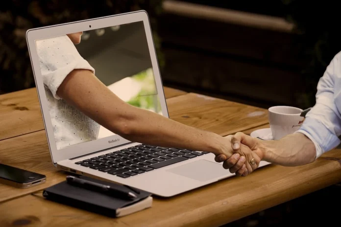 A woman’s arm and hand reaching out of a laptop screen to shake hands with someone sitting in front of it.